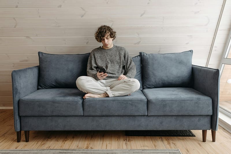 A young man sitting cross-legged on a blue sofa, looking at his phone, in a minimalist wooden-paneled room, possibly experiencing time blindness as he loses track of time.